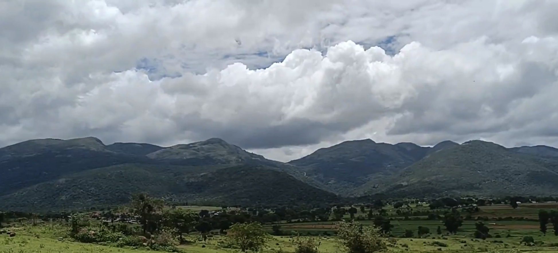 Forested hills rising behind green farmland under monsoon clouds